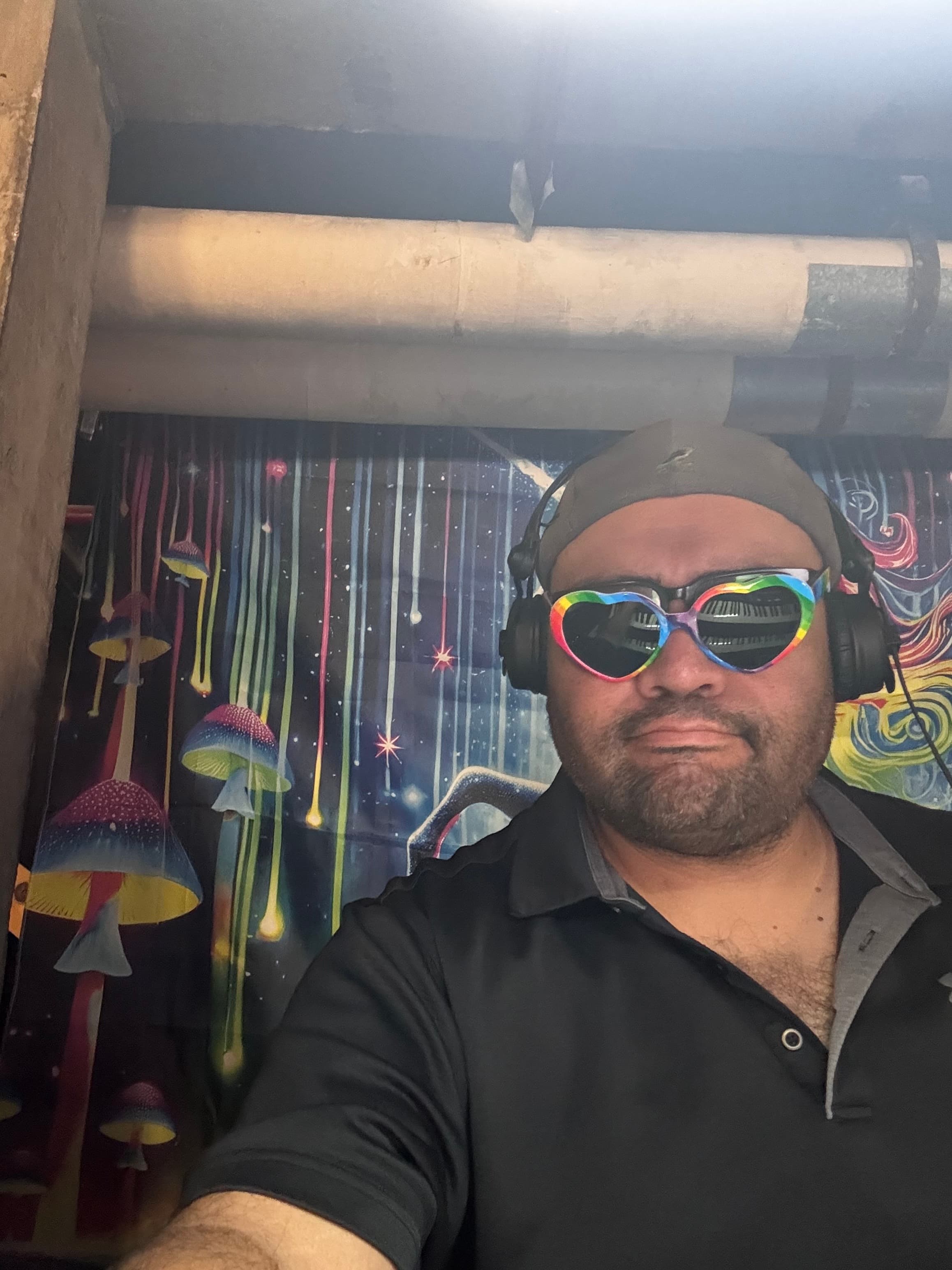 Nathan Spear in rainbow heart sunglasses and headphones in front of a psychedelic mushroom mural.