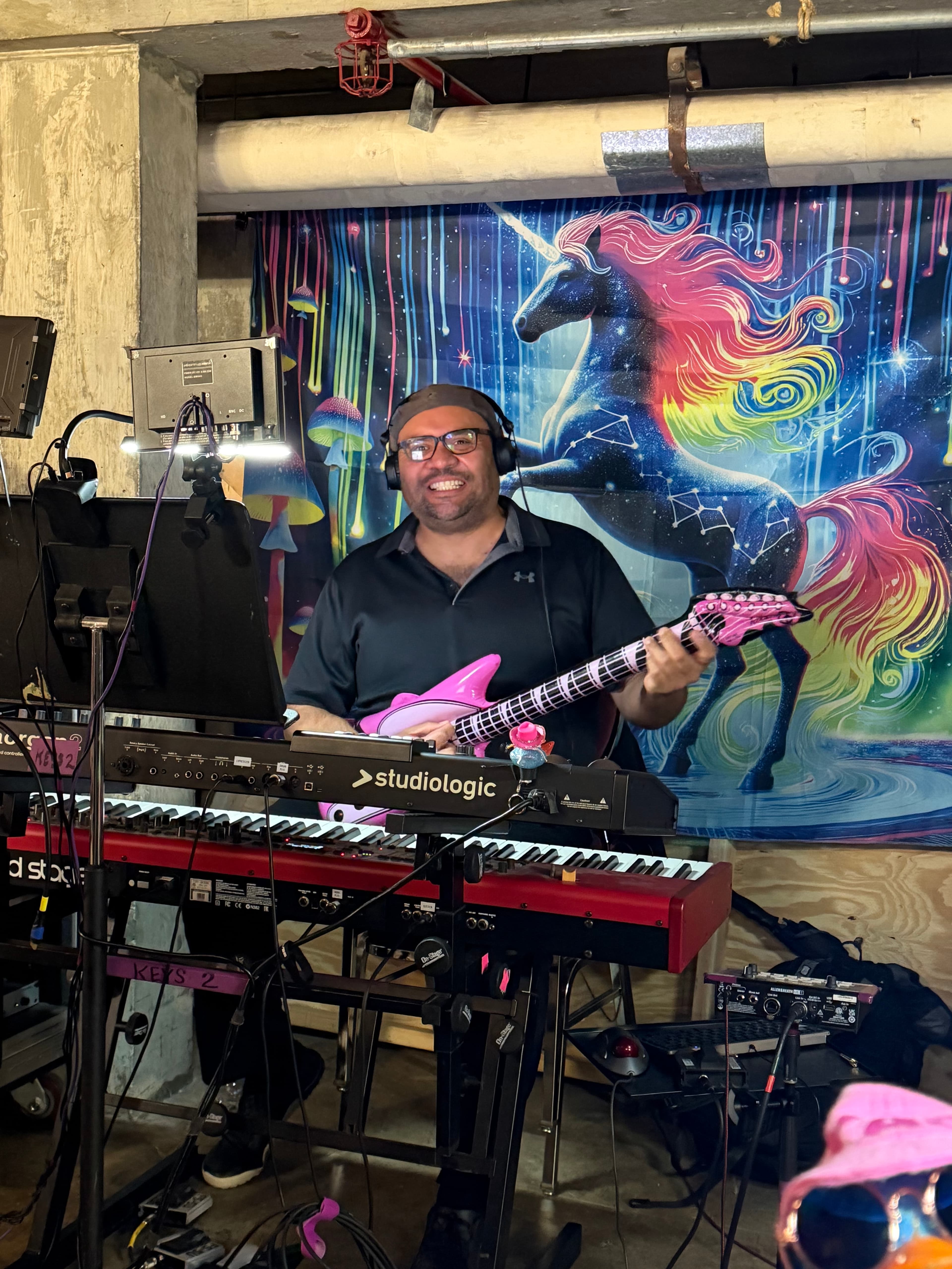Nathan Spear smiling with a pink guitar behind keyboards, a rainbow unicorn backdrop behind him.
